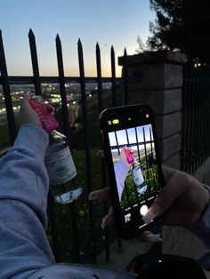 someone taking a photo with their cell phone while standing next to a fence and drinking water