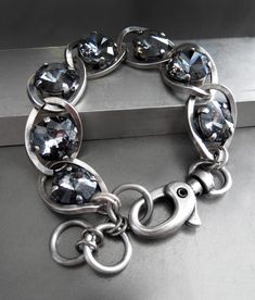 Edgy, sexy, bad-ass. This tough-girl crystal bracelet features exquisite Swarovski crystals in 'Silver Crystal Night' (dark charcoal grey). The sparkling crystals -- alternating both cushion-cut and round rivoli styles -- are interspersed within thick, chunky antiqued silver plated chain links. The length is adjustable from approx. 6" - 7.25" to with the oversized oval lobster clasps and three large rings. Swarovski rivoli crystals: 1/2" dia (12mm dia) Swarovski cushion-cut crystals: 1/2" x 1/2" Large Rings, Black Leather Moto Jacket, Black Lipstick, Black Night, Swarovski Crystal Bracelet, Chain Links, Black Bracelets, Large Ring, Midnight Black