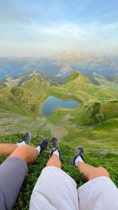 two people sitting on top of a grass covered hill with mountains in the back ground