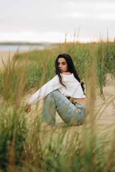 a woman is sitting in the sand by some tall grass and looking at the camera