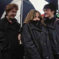 three people standing under an umbrella in the rain, one is smiling at the camera