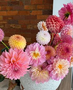 a white vase filled with lots of pink and yellow flowers next to a brick wall