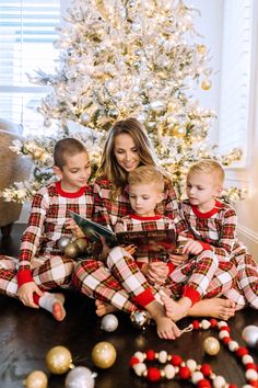 a woman sitting on the floor with two children in matching pajamas reading a book next to a christmas tree