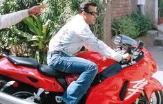 two men sitting on the back of a red motorcycle talking to each other in front of some bushes