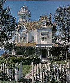 an old house with a white picket fence around it and a light house in the background