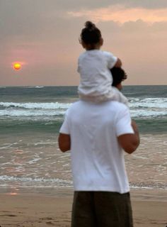 a man holding a child on his shoulders while standing in front of the ocean at sunset