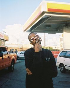 a man standing in front of a gas station talking on a cell phone while wearing sunglasses