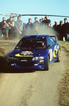 a blue car driving down a dirt road next to a group of people and a helicopter