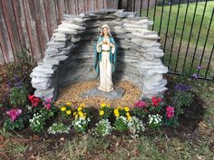 a statue of the virgin mary in front of a fence with flowers and rocks around it