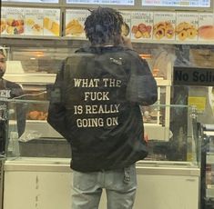 a man standing in front of a donut shop looking at his cell phone while wearing a jacket that says what the fock is really going on