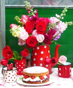 a red vase filled with lots of flowers sitting on top of a table next to a cake