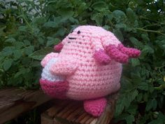 a pink crocheted stuffed animal sitting on top of a wooden bench next to green leaves