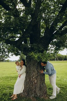 a man and woman standing next to a tree with a baby in their arms on the ground