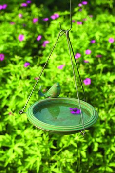 a green bird feeder hanging from a wire with purple flowers in the back ground behind it