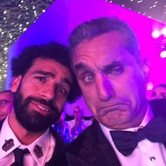 two men in tuxedos pose for a photo at an event with people looking on