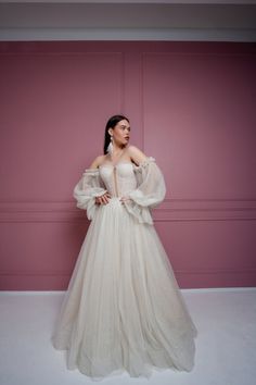 a woman in a white dress standing against a pink wall with her hands on her hips