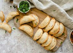 several empanadas on a cutting board next to bowls of sauce and herbs