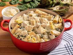 a red pot filled with pasta and vegetables on top of a wooden table next to other ingredients