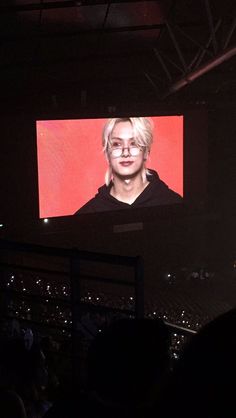 a person with glasses is on a large screen in a dark room while others watch