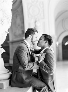 two men in suits are sitting on a bench and one is kissing the other man