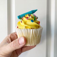 a hand holding a cupcake with yellow frosting and sprinkles