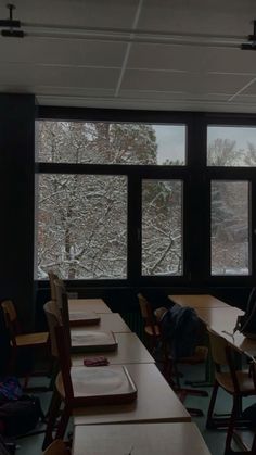 an empty classroom filled with desks and windows covered in snow, looking out onto the trees outside