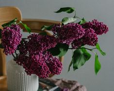 a white vase filled with purple flowers on top of a table next to a wooden chair