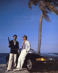 two men standing next to a car on the beach with palm trees in the background