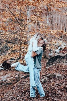 a man and woman hugging in the woods with autumn leaves on the ground behind them