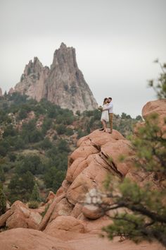 two people standing on top of a rock with trees in the foreground and mountains in the background