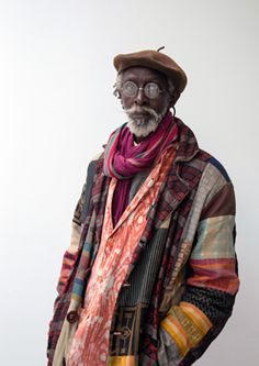 an old man with glasses and a hat is standing in front of a white wall