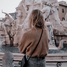 a woman standing in front of a fountain with statues on the sides and buildings behind her