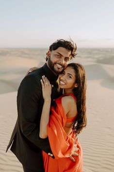 a man and woman in an orange dress embracing each other while standing on sand dunes