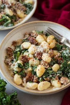 two white bowls filled with pasta and greens on top of a red table cloth next to silverware