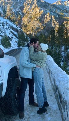 two people standing next to each other in front of a car with mountains in the background