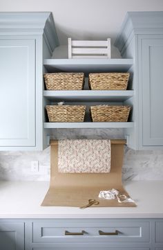 a kitchen with blue cabinets and baskets on the shelf above the countertop, next to a roll of toilet paper that is wrapped in brown paper