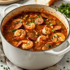 a white pot filled with shrimp and sausage soup on top of a table next to bread