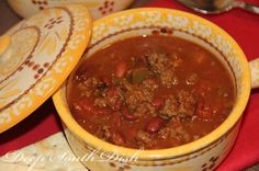 a yellow bowl filled with chili and meat next to a red table cloth on top of a wooden table