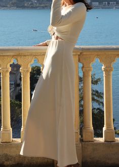 a woman in a long white dress standing on a balcony looking at the water and buildings