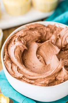 a white bowl filled with chocolate frosting on top of a blue cloth next to cupcakes