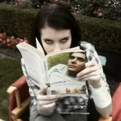 a woman sitting in a chair holding up a book to her face and looking at the camera