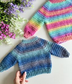 two knitted sweaters sitting on top of a white table next to purple flowers