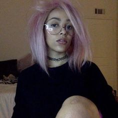 a woman with pink hair and glasses sitting on a bed