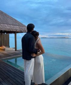 a man and woman standing next to each other on a dock by the water at dusk