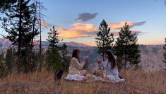 two women sitting on top of a grass covered hill next to each other in front of trees