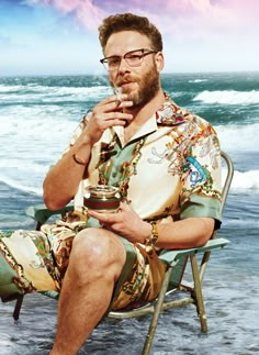 a man sitting on top of a chair next to the ocean holding a glass in his hand