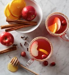 two glasses of apple cider next to cinnamon sticks and orange slices on a marble table