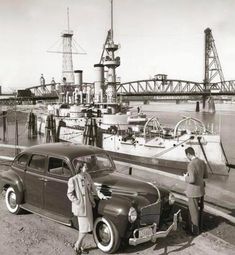 two men standing next to an old car in front of a boat