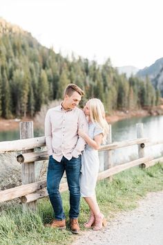 an engaged couple standing next to a wooden fence by the water in front of some pine trees