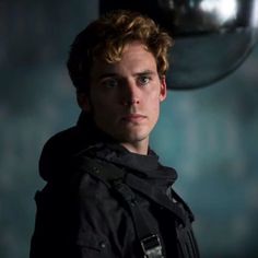 a young man with curly hair wearing a black jacket and looking at the camera while standing in a dark room